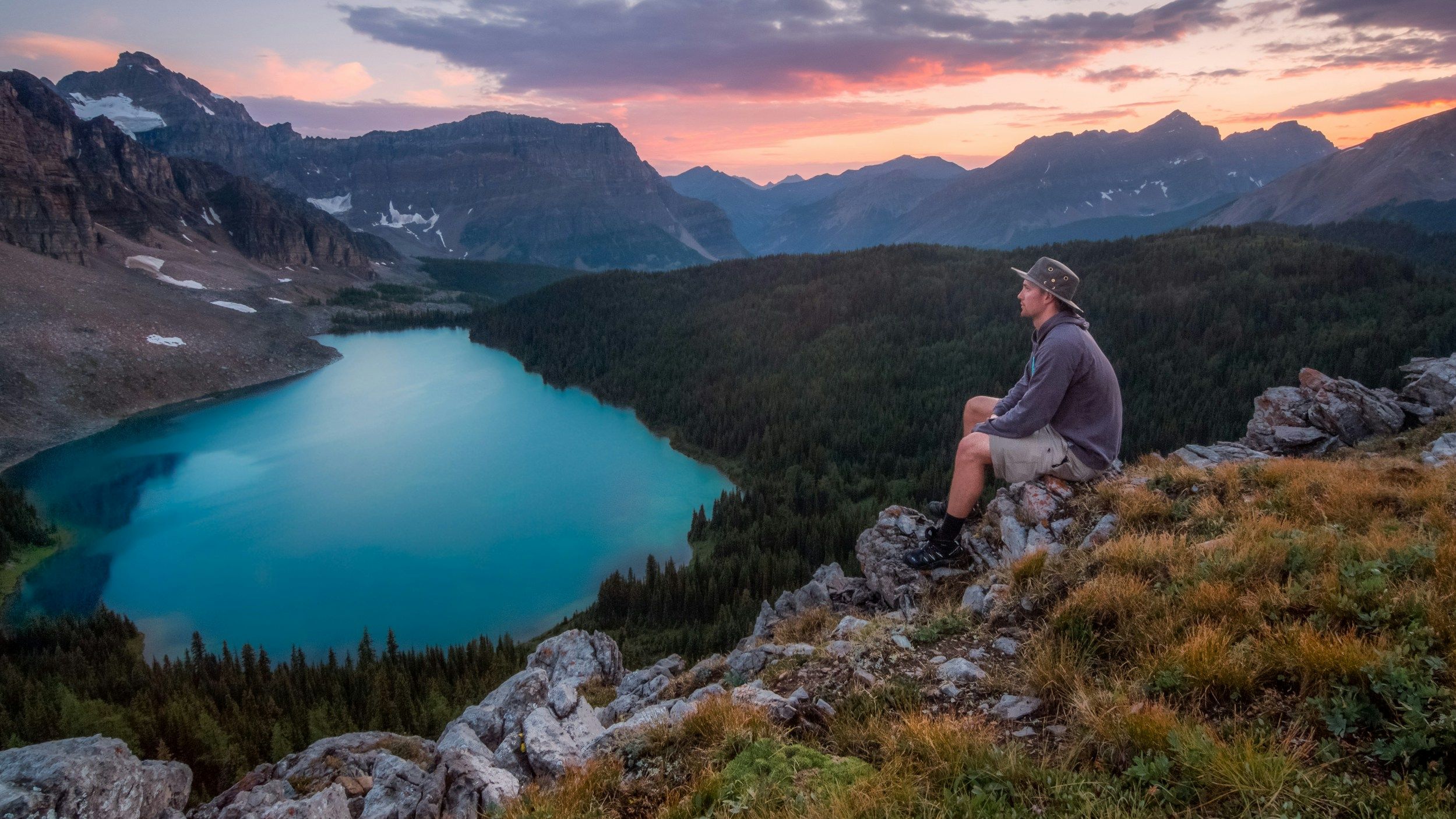 A man sitting on some rocks overlooking a lake surrounded by forests and mountains